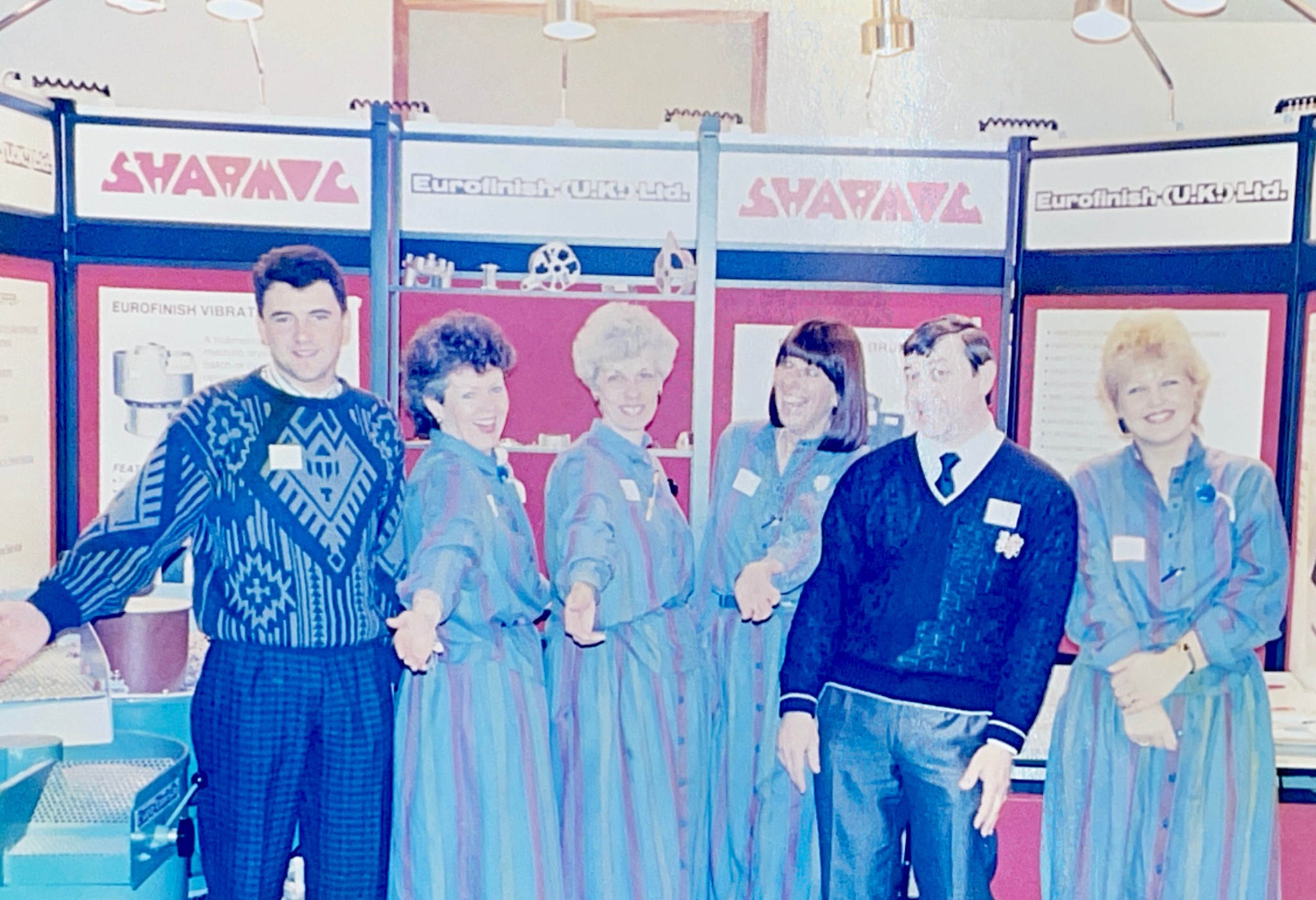 Retired Sharmic Team Members at Exhibition Six former Sharmic Engineering team members, now retired, smiling together at an exhibition with the Sharmic logo and company information displayed in the background.