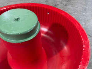 Close-up interior view of a red polyurethane-lined vibratory bowl, showing the ribbed inner wall and central green-painted column with a hex bolt, part of an old industrial vibratory finishing machine.